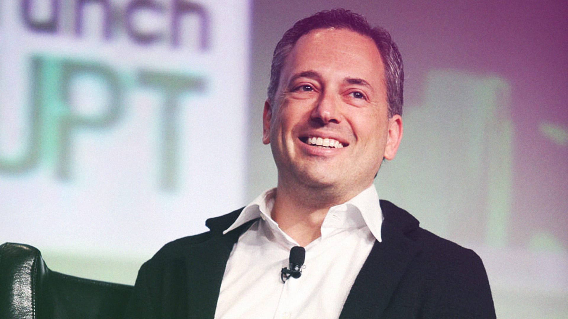 David Sacks smiling while speaking at TechCrunch Disrupt conference, wearing a black blazer and white shirt with a lapel microphone