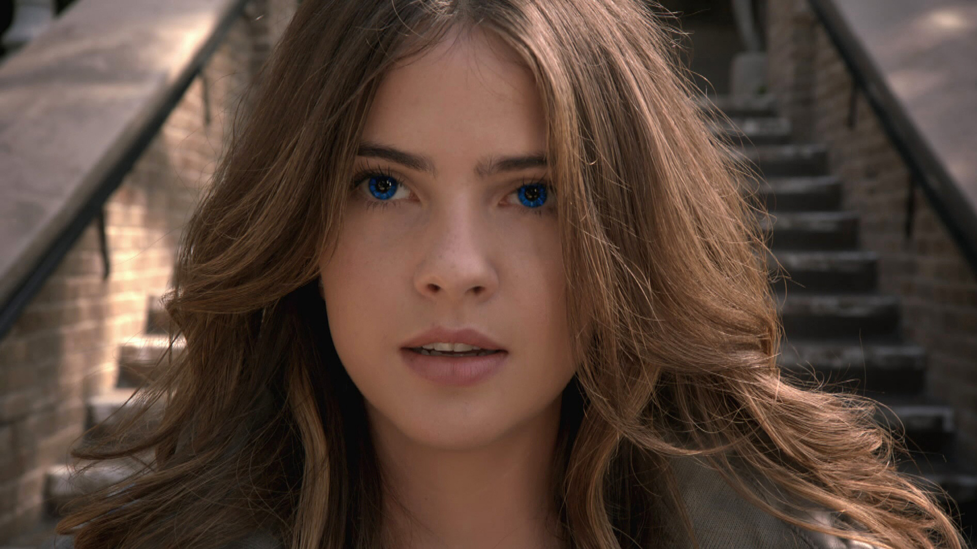 Malia Manocherian close-up portrait with blue eyes and flowing brown hair outdoors near stone steps