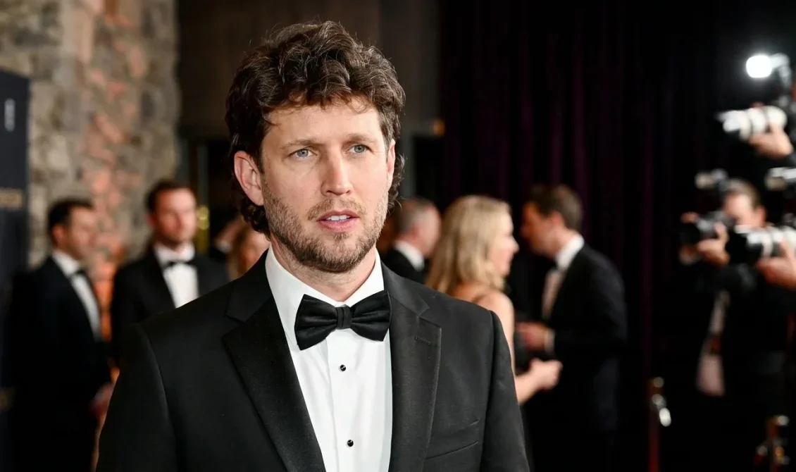 Jon Heder wearing a black tuxedo and bow tie at a formal Hollywood red carpet event with photographers in the background.