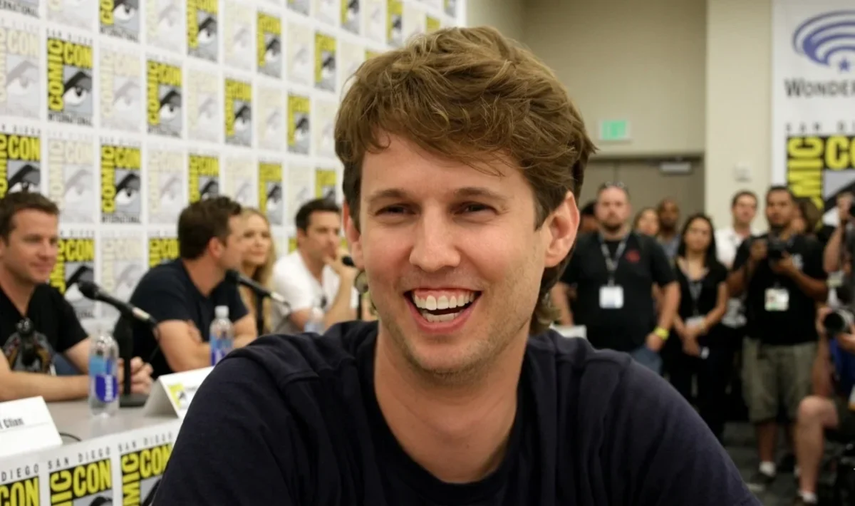 Jon Heder smiling and laughing during a panel session at San Diego Comic-Con, with photographers and fans visible in the background.