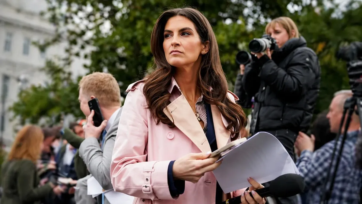 A focused brunette female news Kaitlan Collins Married correspondent in a pink trench coat holding documents and notes while reporting outdoors at a press event near a government building, with photographers and media personnel visible in the background.