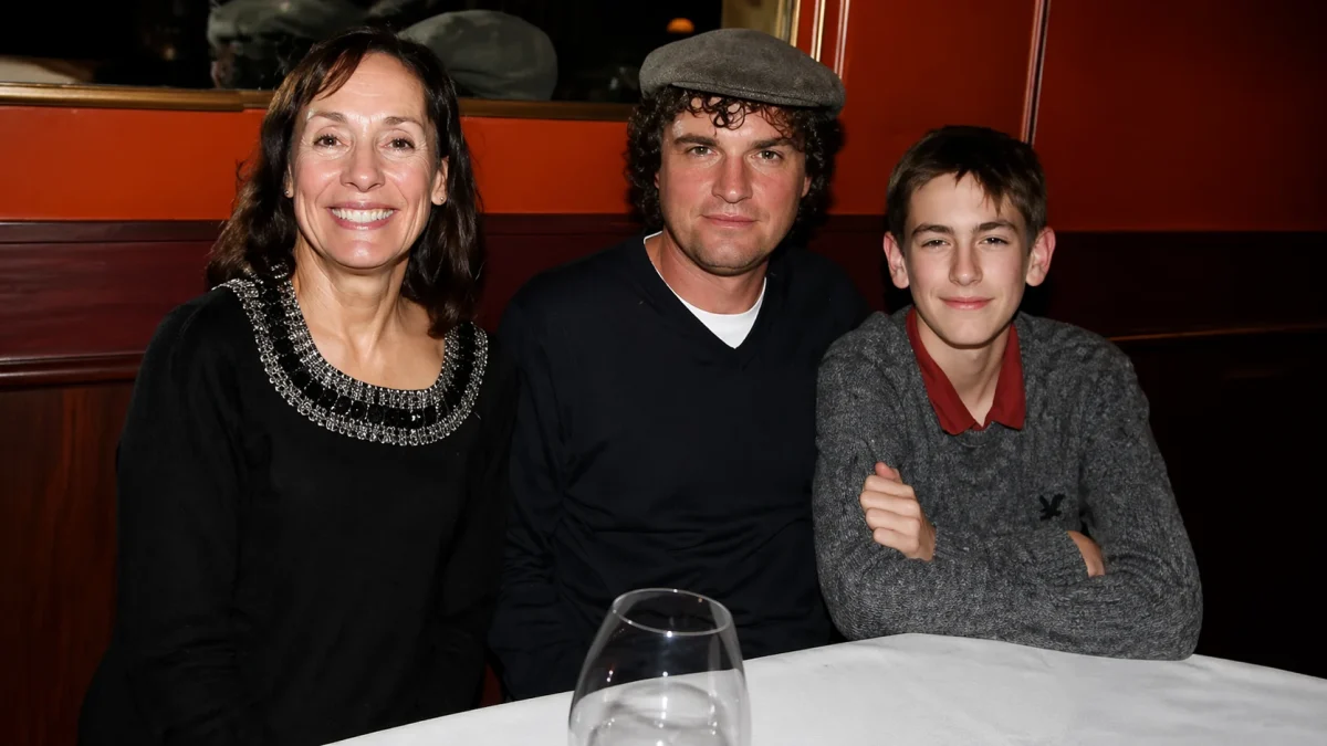 A teenage boy Will Theron Roth with arms crossed seated at a restaurant dinner table alongside a smiling woman in a black dress with a statement necklace and a man in a black sweater and grey flat cap, capturing a warm candid family moment at a private event.