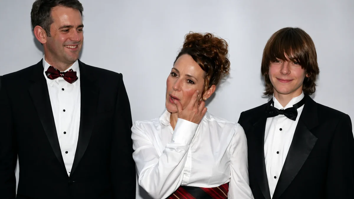 A teenage boy Will Theron Roth in a black tuxedo and bow tie standing alongside a man in a tuxedo with a burgundy bow tie and a woman in a white blouse with an updo hairstyle at a formal Hollywood gala event against a white backdrop.