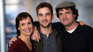 A young adult man Will Theron Roth with hazel eyes and light stubble smiling with his arms around a brunette woman in a burgundy top and a curly-haired man in a grey flat cap and black sweater, posing together for a warm and close family portrait against a softly blurred modern background.