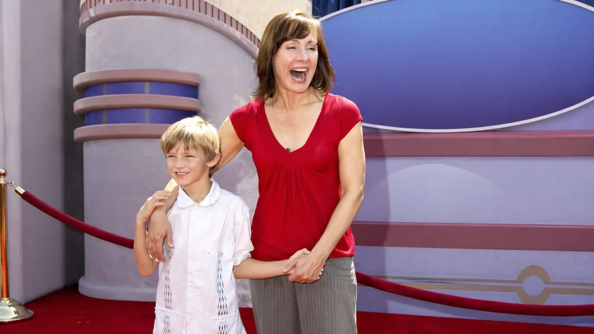A young blonde boy will theron roth smiling alongside a laughing brunette woman in a red top at a Hollywood red carpet premiere event, posing together in front of a colorful theatrical backdrop behind a velvet rope.