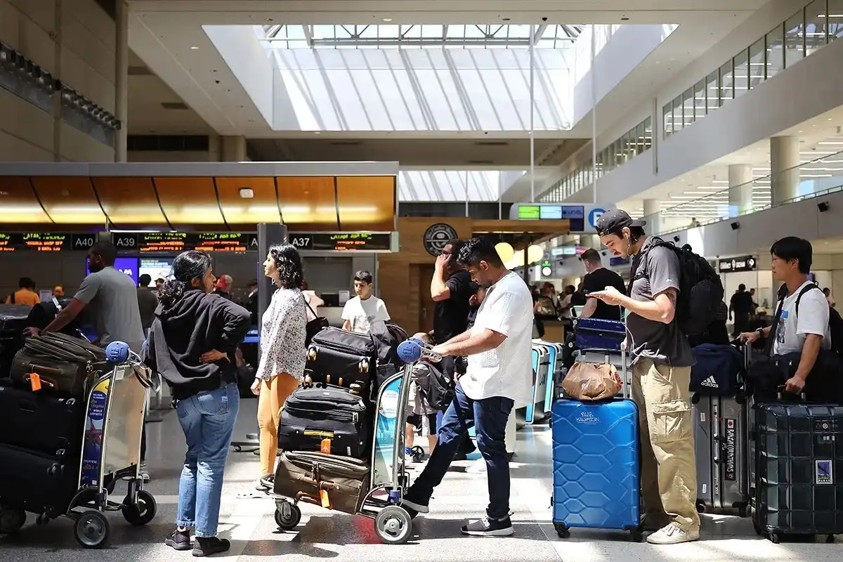 A busy Delta Air Lines airport terminal check-in area with diverse travelers standing in line with luggage carts piled with suitcases, blue and black bags visible near gates A37, A39, and A40, under a bright skylight ceiling in a modern terminal building.