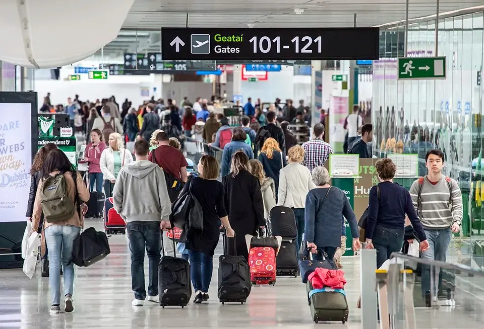 A large crowd of travelers with rolling suitcases and backpacks walking through a busy modern airport terminal toward gates 101 to 121, with bilingual Irish and English signage visible overhead in what appears to be Delta Airport's departure hall.