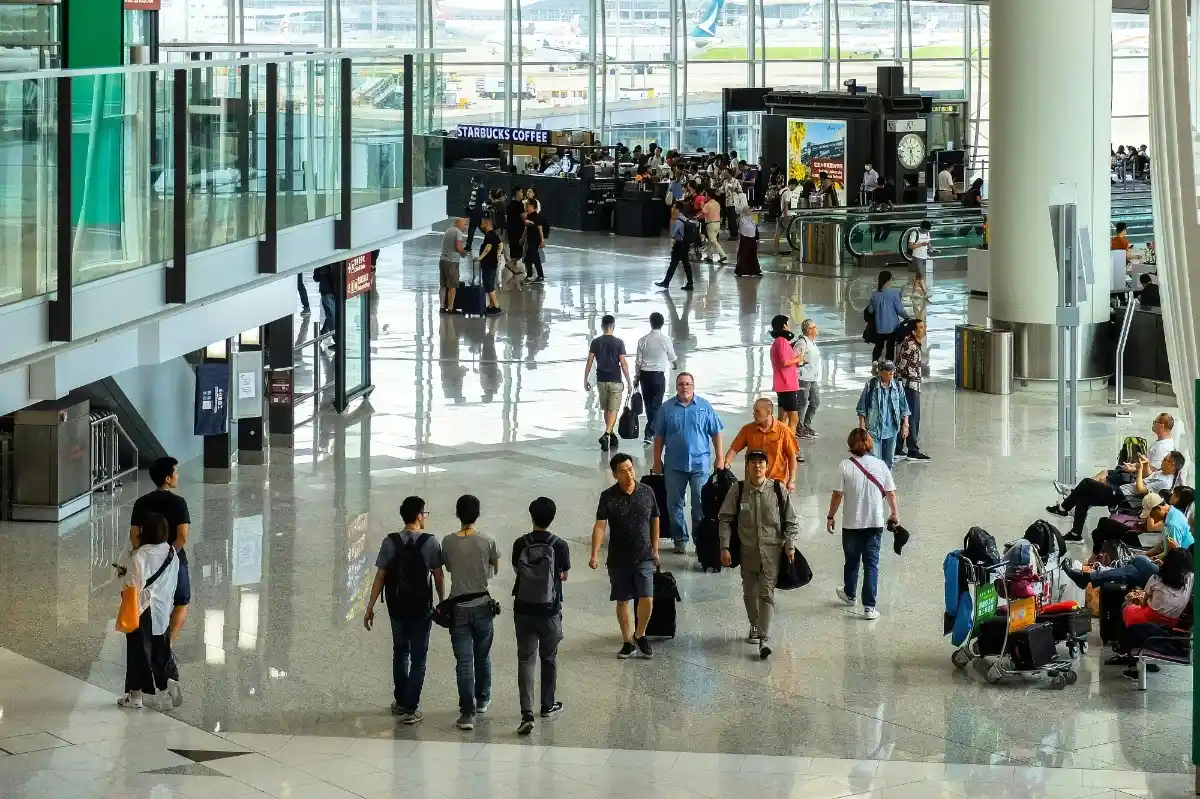 A long queue of diverse travelers with colorful rolling suitcases and backpacks standing in line at a busy delta airport terminal boarding gate, with departure information boards visible in the blurred background.