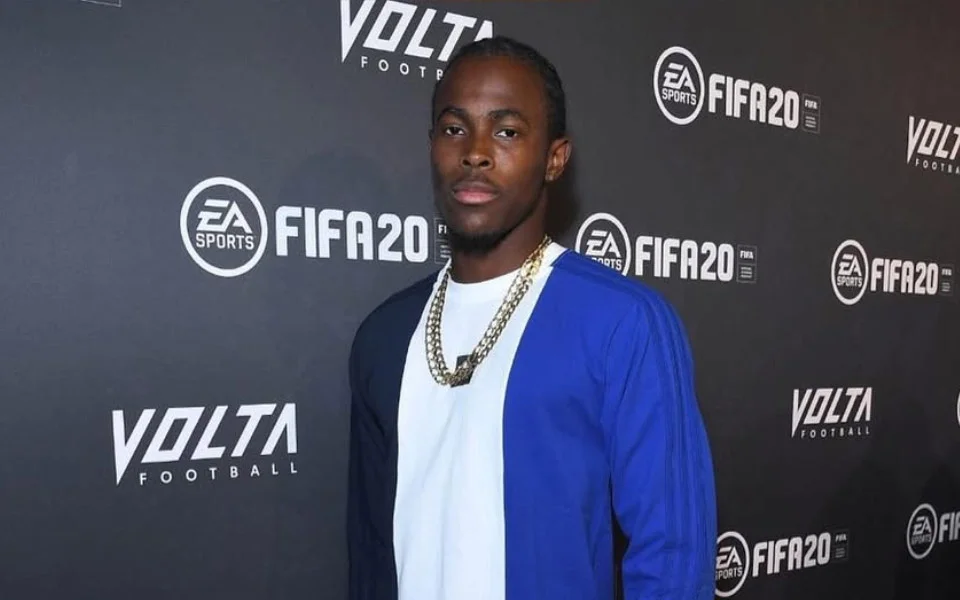 Jofra Archer net worth posing in a blue and white color-block jacket and gold chain necklace at the EA Sports FIFA 20 and Volta Football launch event, standing confidently in front of the branded step-and-repeat backdrop.