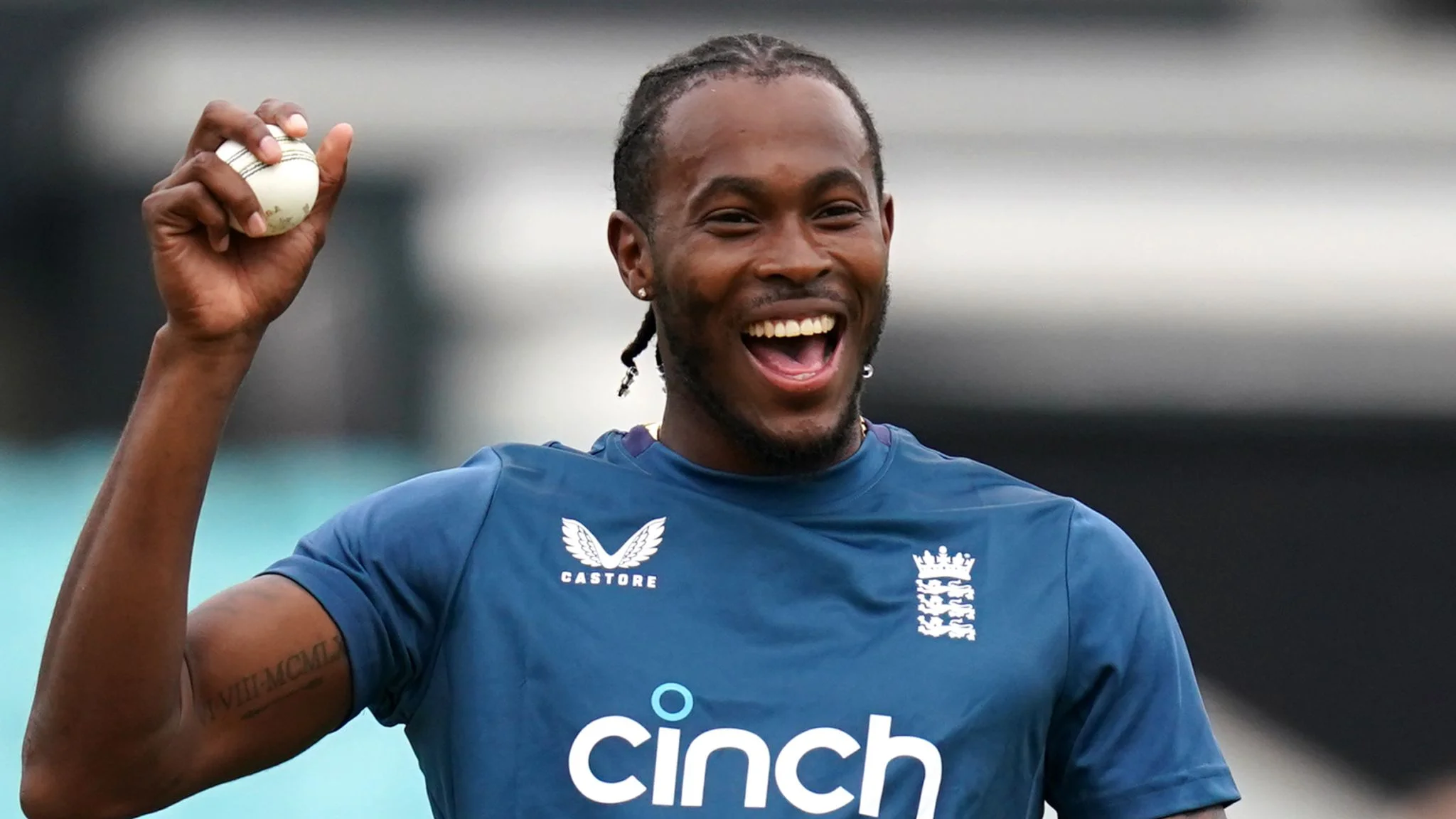 Jofra Archer net worth laughing joyfully while holding a white cricket ball aloft during an England Cricket training session, wearing a navy blue Castore kit sponsored by Cinch with the Three Lions England Cricket badge visible on his chest.
