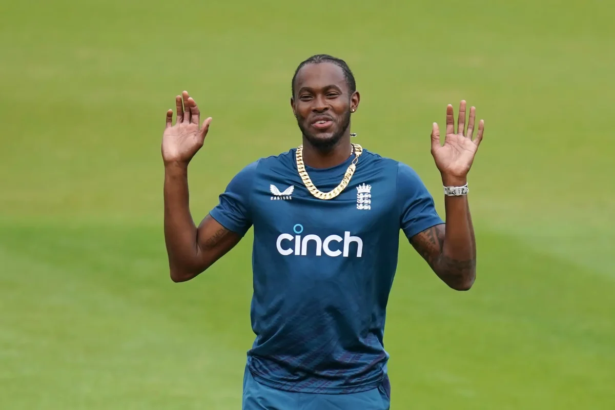 Jofra Archer net worth smiling with both hands raised on a green cricket outfield, wearing an England Cricket Castore training kit sponsored by Cinch, a gold chain necklace, and a silver bracelet, reflecting his confident and stylish personality.