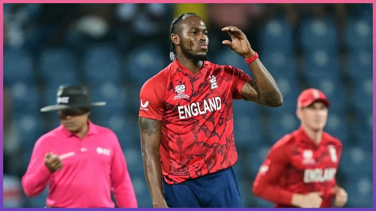 Jofra Archer net worth in England's red T20 World Cup kit gesturing confidently on the cricket field under floodlights, with an umpire in pink and a teammate visible in the background during an international match.