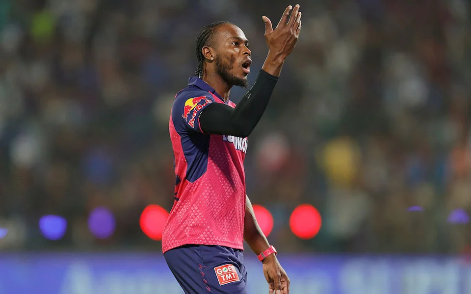 Jofra Archer net worth celebrating an emotional wicket in his pink Rajasthan Royals IPL kit with Red Bull and Liquigas sponsorship logos, raising his hands expressively under floodlights during an Indian Premier League match in front of a packed crowd.