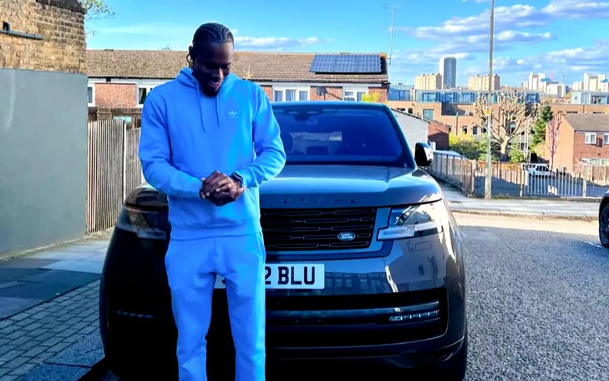Jofra Archer net worth wearing a light blue Adidas tracksuit smiling while standing in front of a dark grey Range Rover with a personalised "2 BLU" number plate on a residential London street on a sunny day.