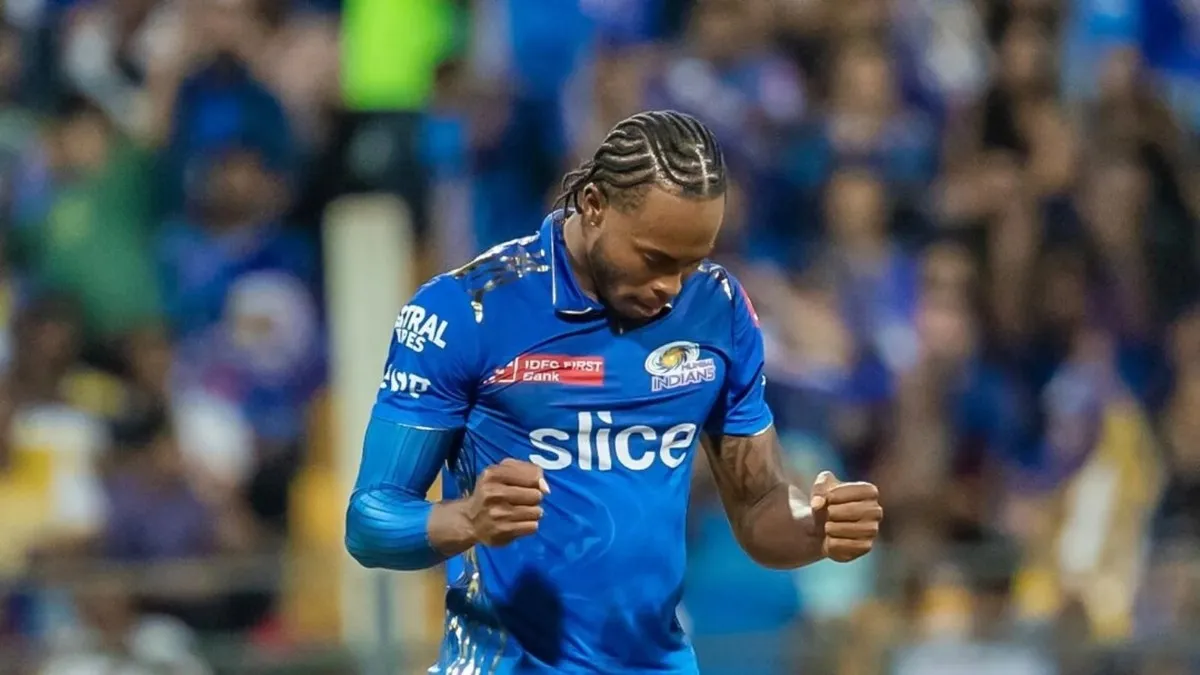 Jofra Archer net worth pumping his fists in an intense celebratory gesture wearing a blue Mumbai Indians IPL kit sponsored by Slice and IDFC First Bank, with braided hair and a packed stadium crowd visible in the blurred background.