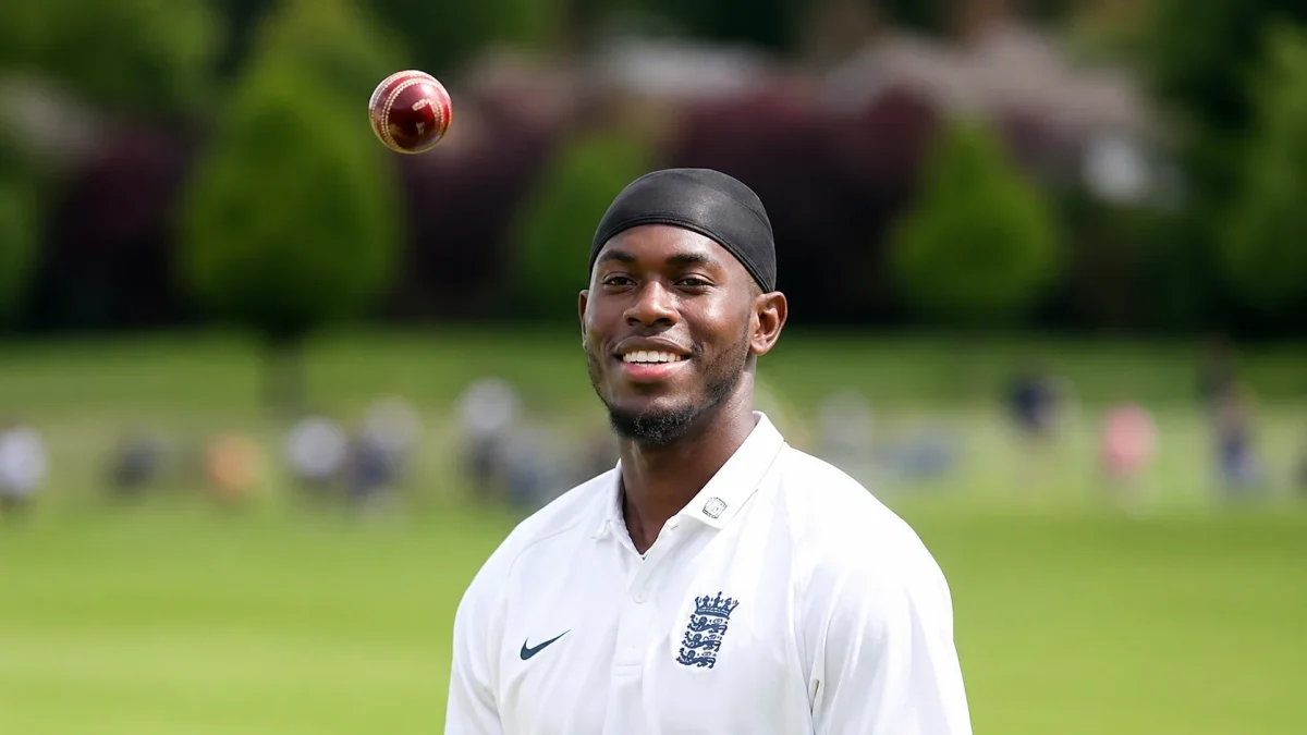 A smiling young male cricket player wearing a white Sussex Cricket kit sponsored by Pembroke, tossing a red cricket ball in the air on a green cricket ground, representing England pace bowler Jofra Archer net worth.