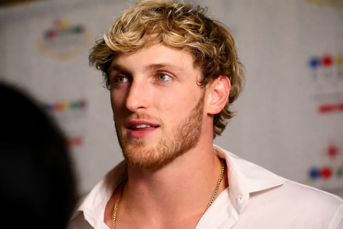 A close-up portrait of Logan Paul with curly blonde hair and green eyes wearing a white open-collar shirt and gold chain necklace at a press event, capturing the charismatic persona that has built Logan Paul net worth into one of the most impressive in digital entertainment history.