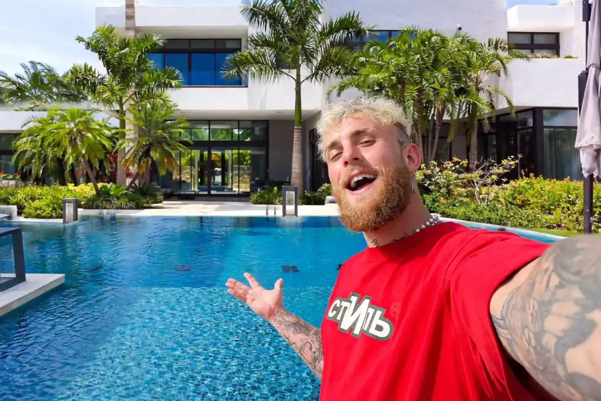 Logan Paul taking an excited selfie in front of a stunning luxury mansion with a large infinity pool and tropical palm trees, giving a vivid glimpse into the lavish lifestyle funded by Logan Paul net worth from his YouTube, WWE, boxing, and Prime Hydration empire.