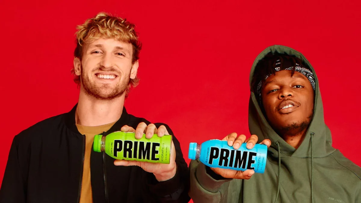 Logan Paul and KSI smiling together against a bold red background while holding green lemon lime and blue raspberry Prime Hydration bottles — the co-founded beverage brand that dramatically elevated Logan Paul net worth to new heights.