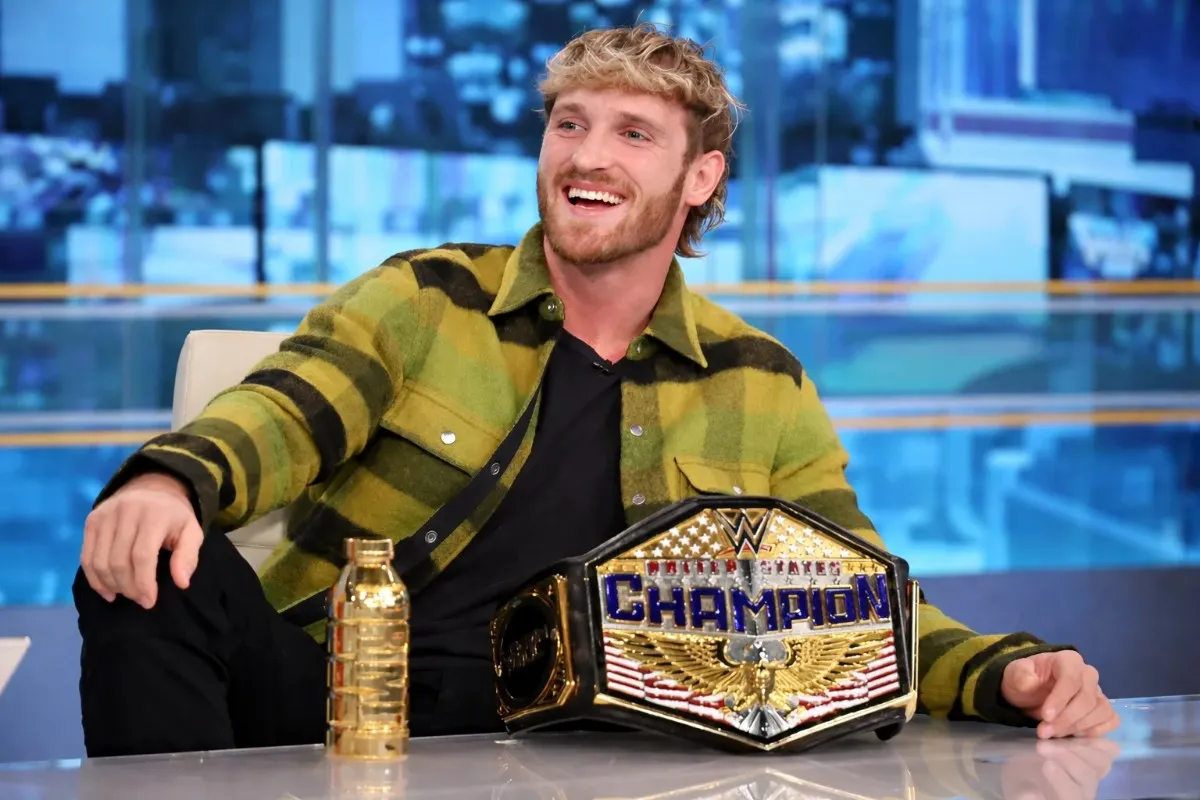 Logan Paul laughing joyfully at a television talk show desk with the WWE United States Championship belt prominently displayed in front of him, showcasing the championship achievement that has contributed significantly to Logan Paul net worth through WWE earnings and media appearances.