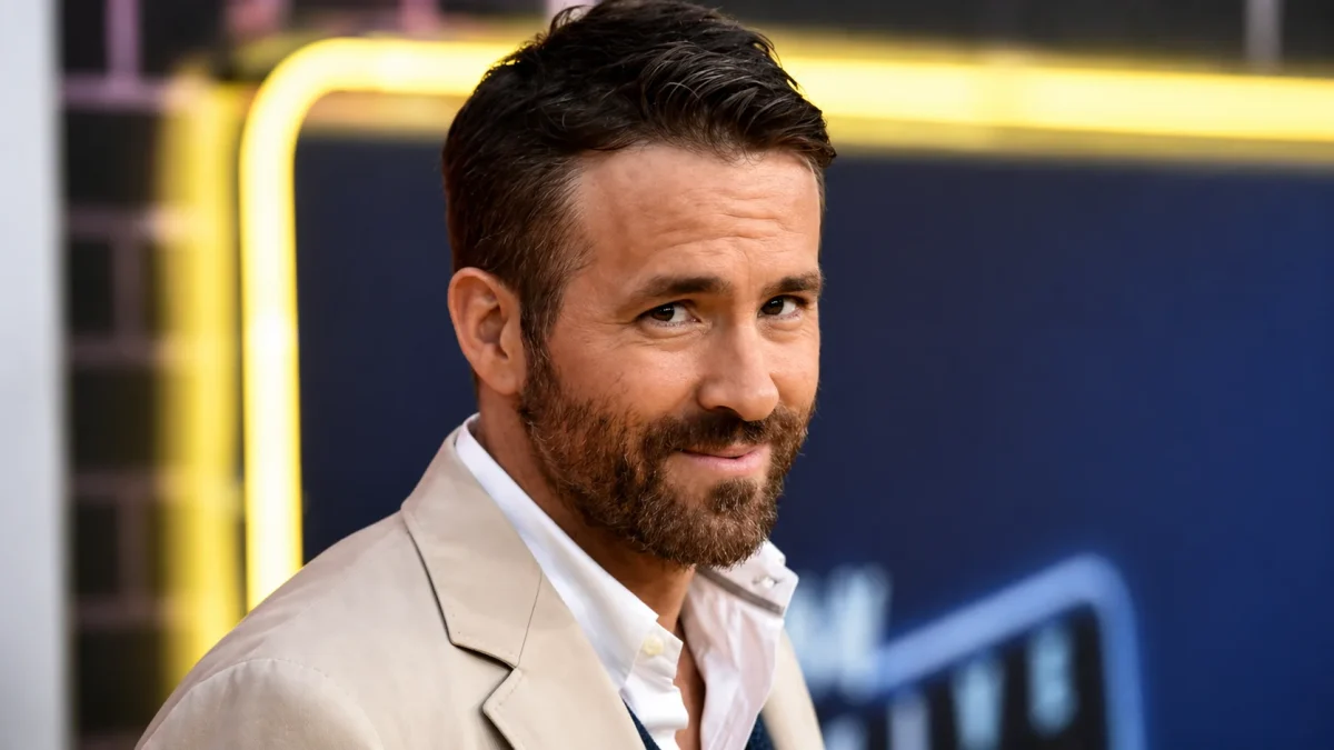 A close-up of Ryan Reynolds giving a confident over-the-shoulder glance at a Hollywood movie premiere wearing a beige blazer and white shirt, perfectly capturing the leading man charisma that has driven Ryan Reynolds net worth to over $350 million.
