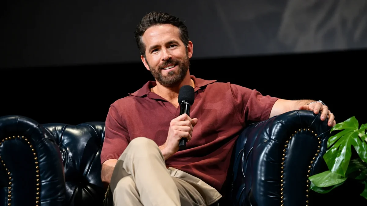 Ryan Reynolds relaxed and smiling on stage holding a microphone while seated in a navy blue leather Chesterfield armchair during a public speaking event, embodying the confident entrepreneurial persona behind Ryan Reynolds net worth success story.