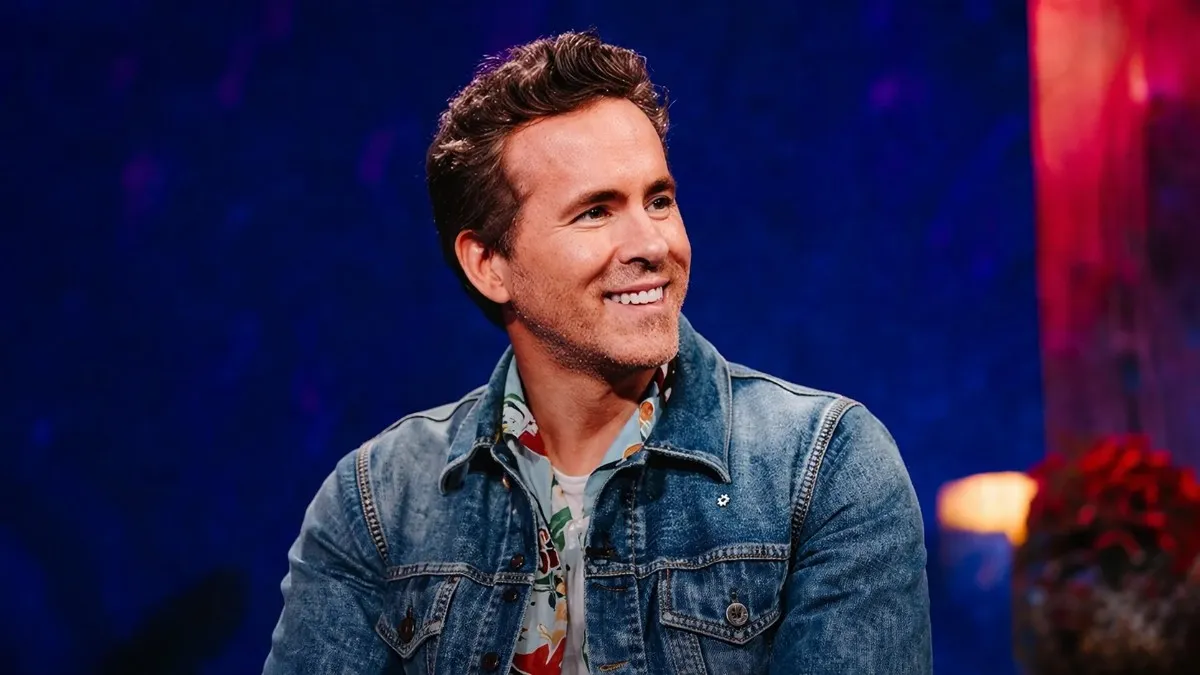 Ryan Reynolds smiling broadly during a television talk show appearance wearing a denim jacket and floral shirt, reflecting the charismatic personality that has helped build Ryan Reynolds net worth through acting, business ventures, and brand partnerships.