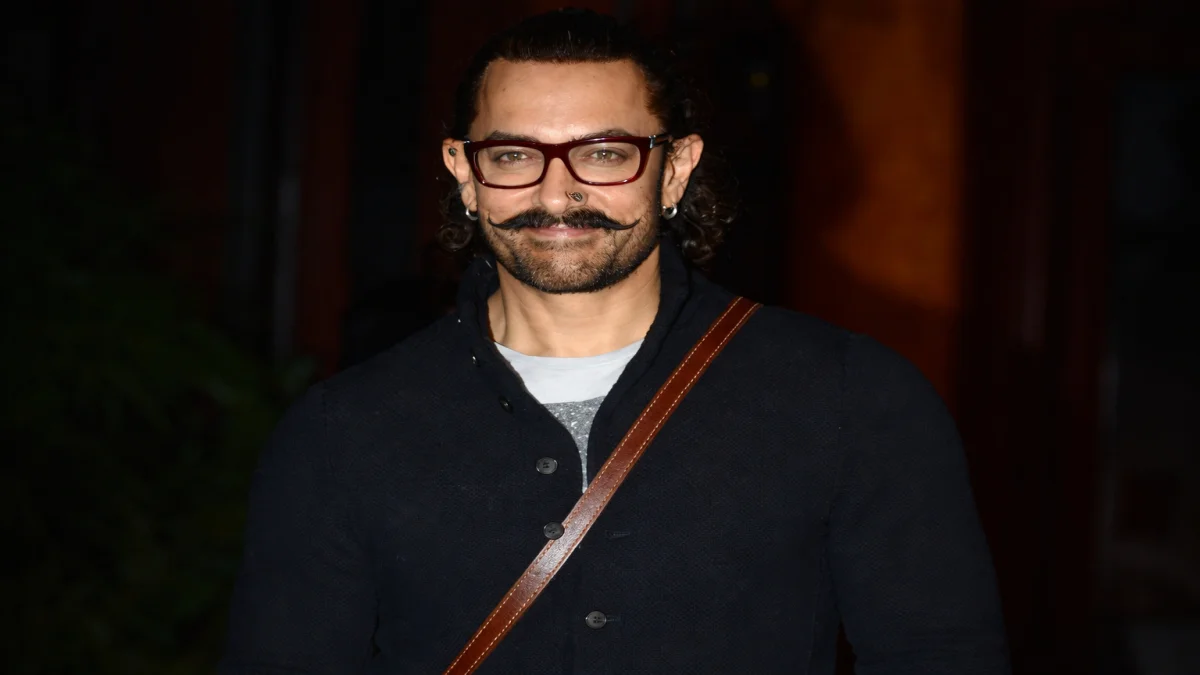 Aamir Khan net worth smiling in red-framed glasses, curly hair, and a navy jacket with a brown leather crossbody bag, reflecting his iconic off-screen personal style