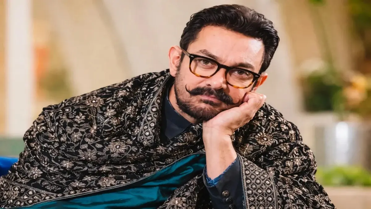 Aamir Khan net worth wearing an ornate black and gold embroidered sherwani with tortoiseshell glasses, resting his chin on his hand in a thoughtful pose at a formal event