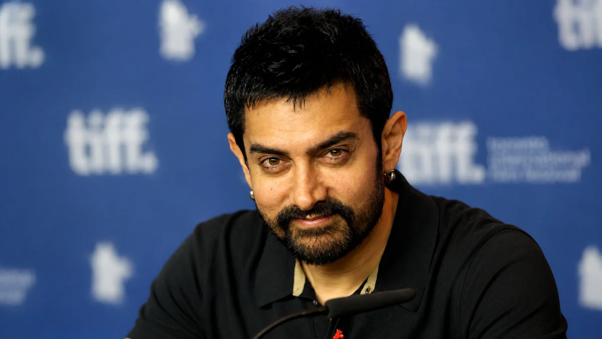 Aamir Khan net worth sitting at a TIFF press conference in a black polo shirt in front of a blue Toronto International Film Festival backdrop