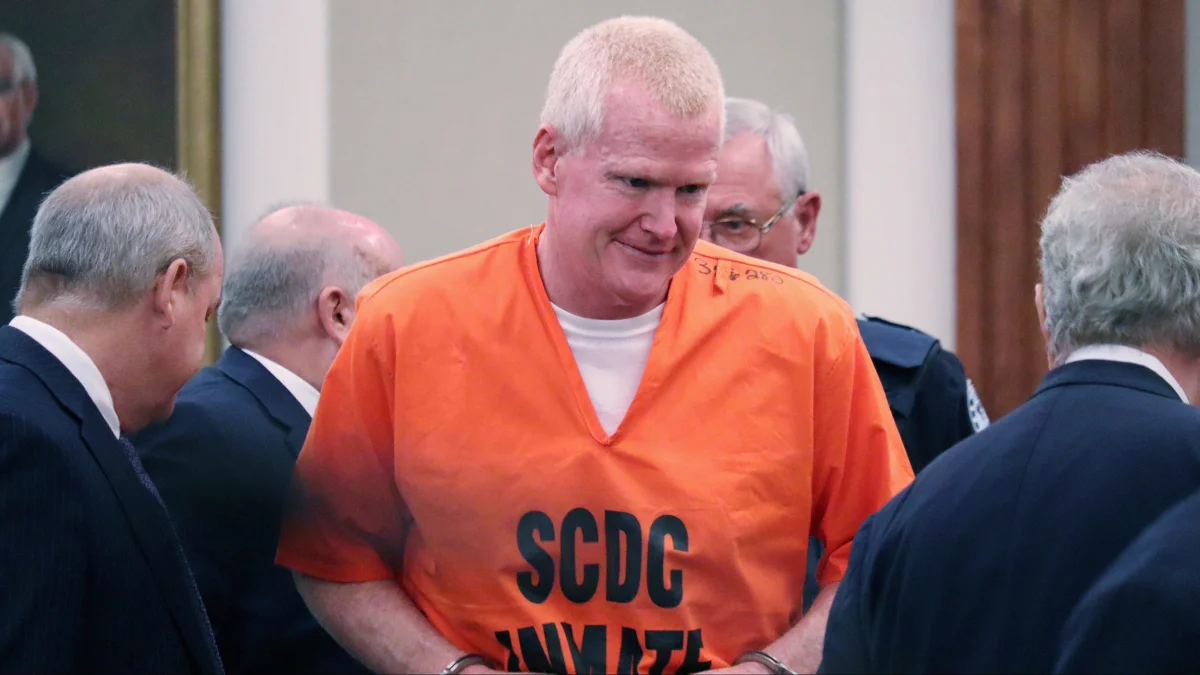 Alex Murdaugh wearing an orange SCDC inmate uniform and handcuffs during a courtroom appearance surrounded by attorneys and law enforcement officers