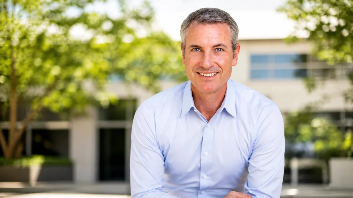 A confident silver-haired male technology CEO with blue eyes smiling warmly in a light blue business shirt outdoors in front of a modern office building surrounded by green trees — representing the polished executive presence and professional leadership that has built Andy Byron net worth through his role at Astronomer.