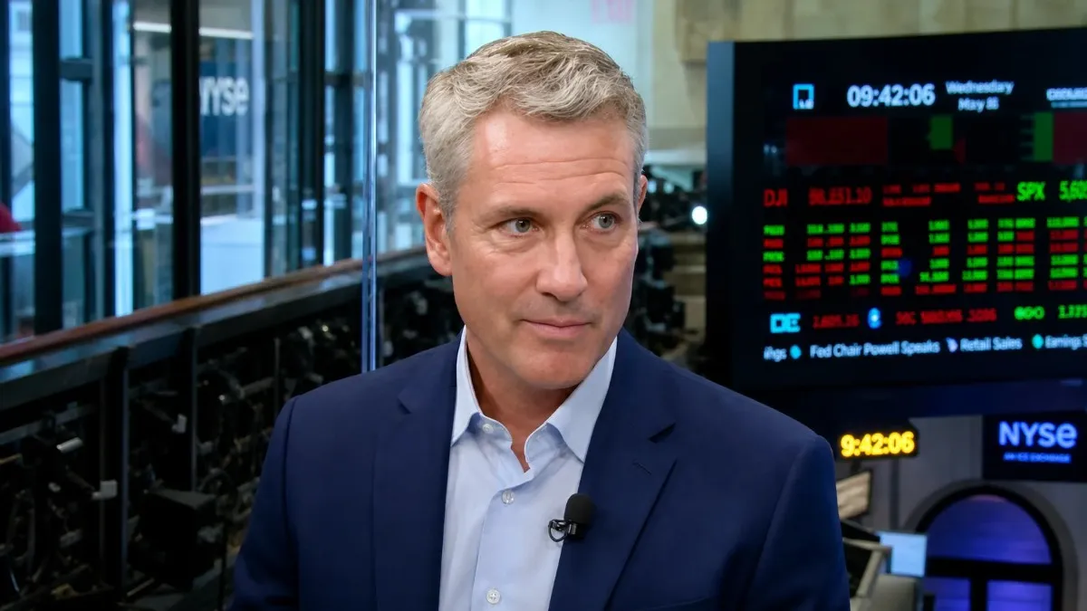 A silver-haired male technology CEO wearing a navy blazer and light blue shirt with a lapel microphone being interviewed at the New York Stock Exchange with live stock ticker screens displaying DJI and SPX market data in the background, representing the high-finance world that shapes Andy Byron net worth as a leading tech executive.