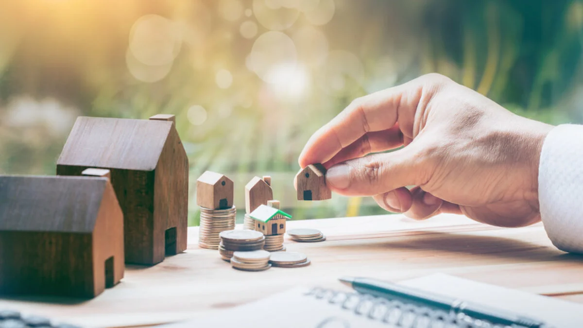 A hand placing a small wooden house model among stacked coins and multiple miniature house figurines on a desk, symbolizing the real estate assets and property investment portfolio that contribute to Andy Byron net worth beyond his tech executive earnings.