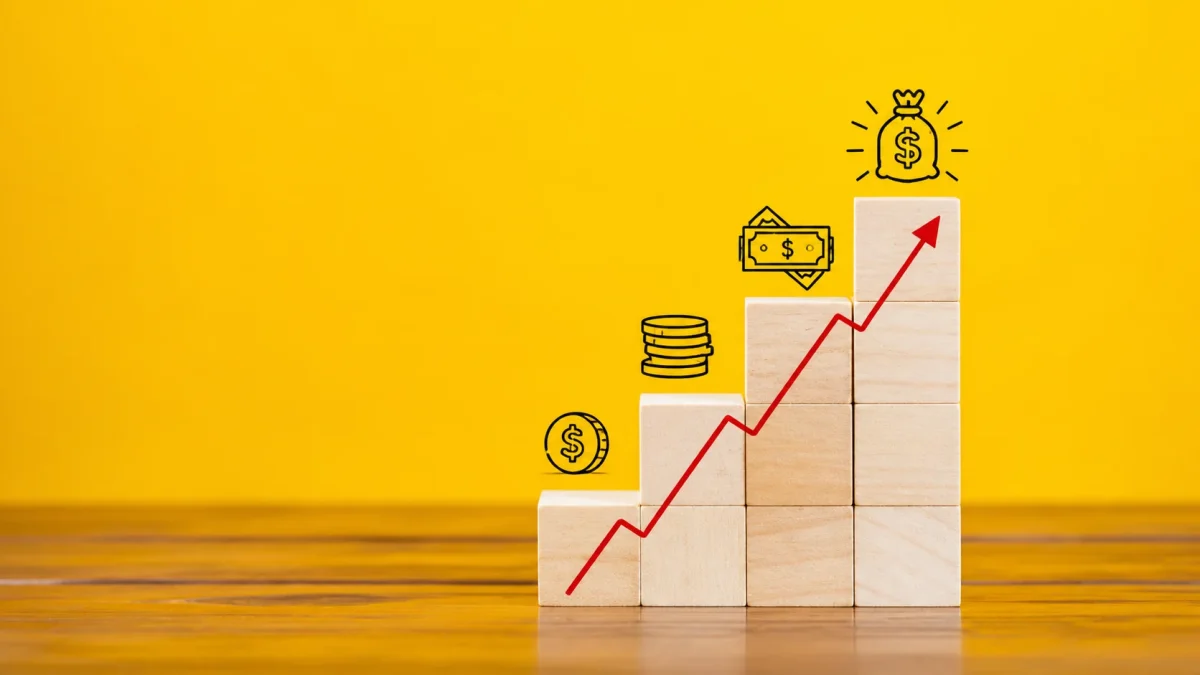 Wooden building blocks arranged in a rising staircase formation with a red upward growth arrow and illustrated financial icons progressing from a single coin to stacked coins, bundled cash, and a glowing money bag at the top — visually representing the step-by-step wealth accumulation journey behind Andy Byron net worth.