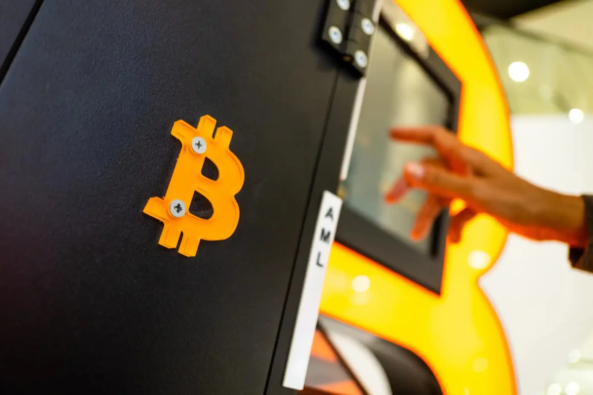 Close-up of a Bitcoin ATM machine with an orange 3D Bitcoin logo mounted on a black panel while a person taps the touchscreen display