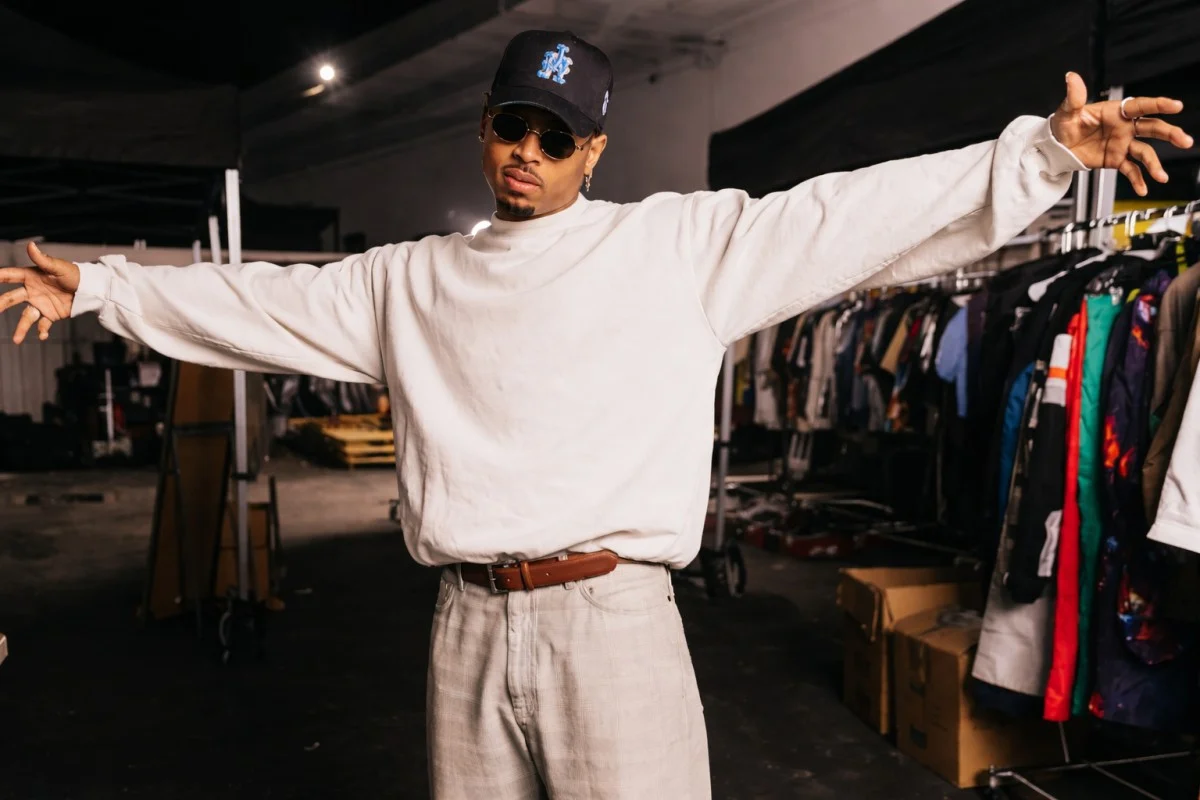 Chris Brown posing with arms stretched wide in a white oversized sweatshirt, light grey trousers, brown leather belt, dark cap, and oval sunglasses in a backstage wardrobe room filled with clothing racks — reflecting the fashion entrepreneurship and personal style that contributes to Chris Brown net worth beyond music.