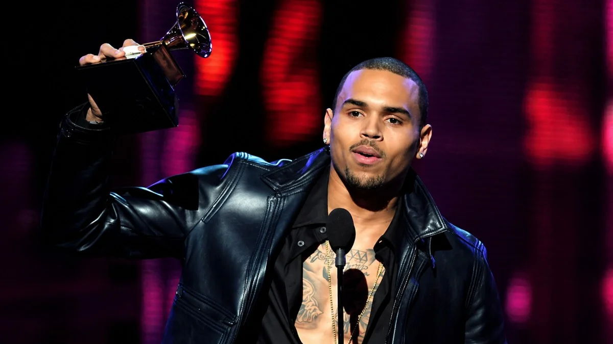  Chris Brown holding a Grammy Award aloft at the microphone podium wearing a black leather jacket with chest tattoos visible and gold chain necklace — a defining career achievement moment that significantly boosted Chris Brown net worth through increased album sales, streaming revenue, and commercial opportunities.