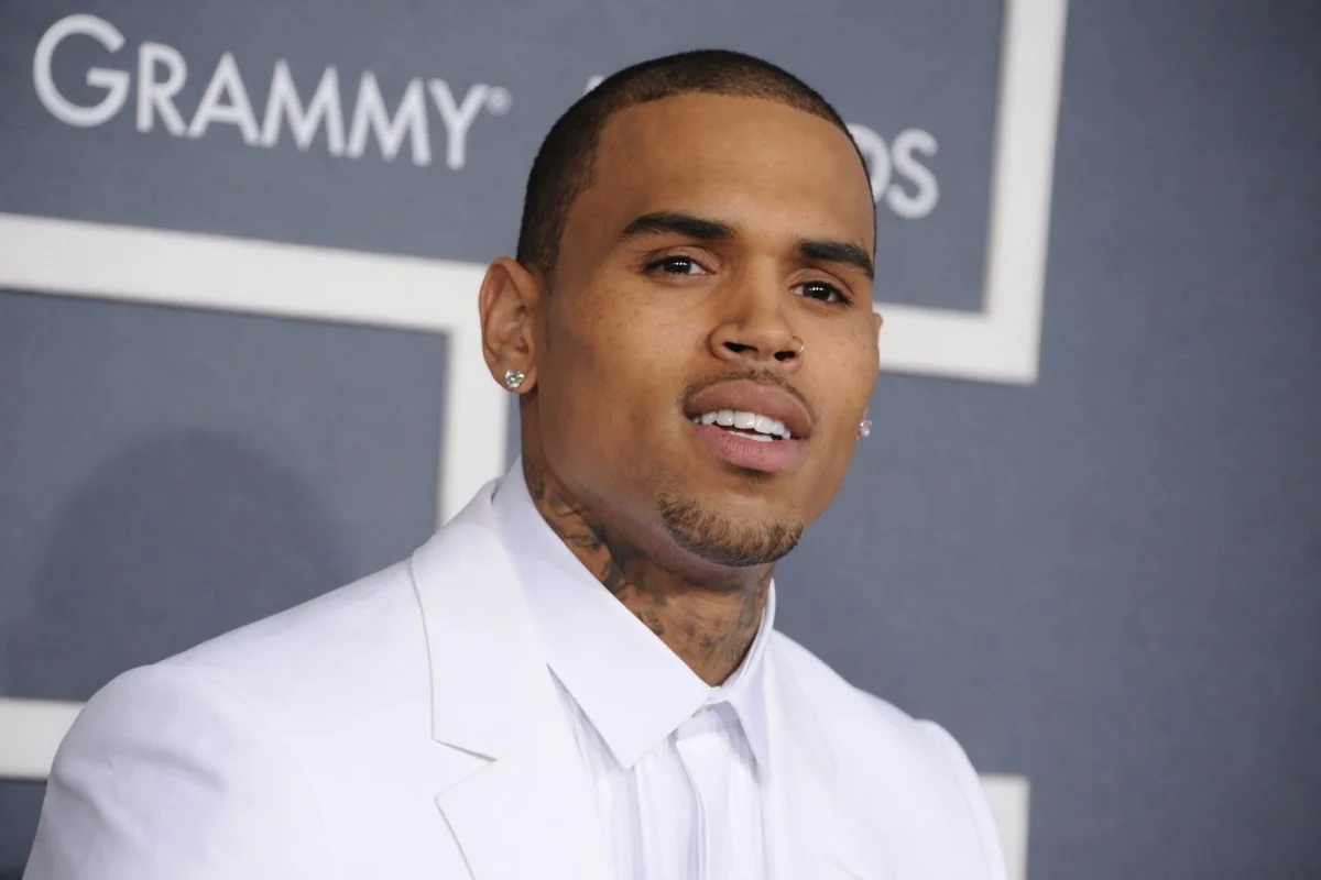 Chris Brown wearing a crisp all-white suit with diamond stud earrings and neck tattoos visible on the Grammy Awards red carpet, projecting the polished elegance and enduring star power that has sustained Chris Brown net worth as one of R&B's most commercially successful artists over two decades.