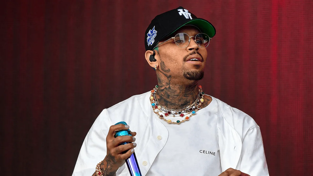 Chris Brown holding a blue microphone on stage wearing a white Celine shirt, black cap, round glasses, layered multicolored gemstone necklaces, and visible neck and arm tattoos — a performance image reflecting the star power and stage presence behind Chris Brown net worth as one of R&B's highest-earning artists.