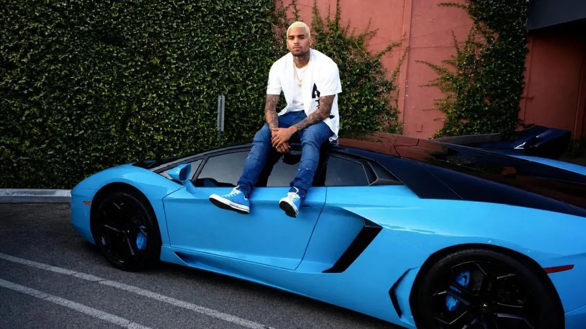  Chris Brown with blonde hair sitting casually on top of a vibrant blue Lamborghini Aventador wearing a white oversized t-shirt, ripped jeans, blue Converse sneakers, and a gold chain — a striking lifestyle image showcasing the luxury assets that reflect Chris Brown net worth and celebrity wealth.