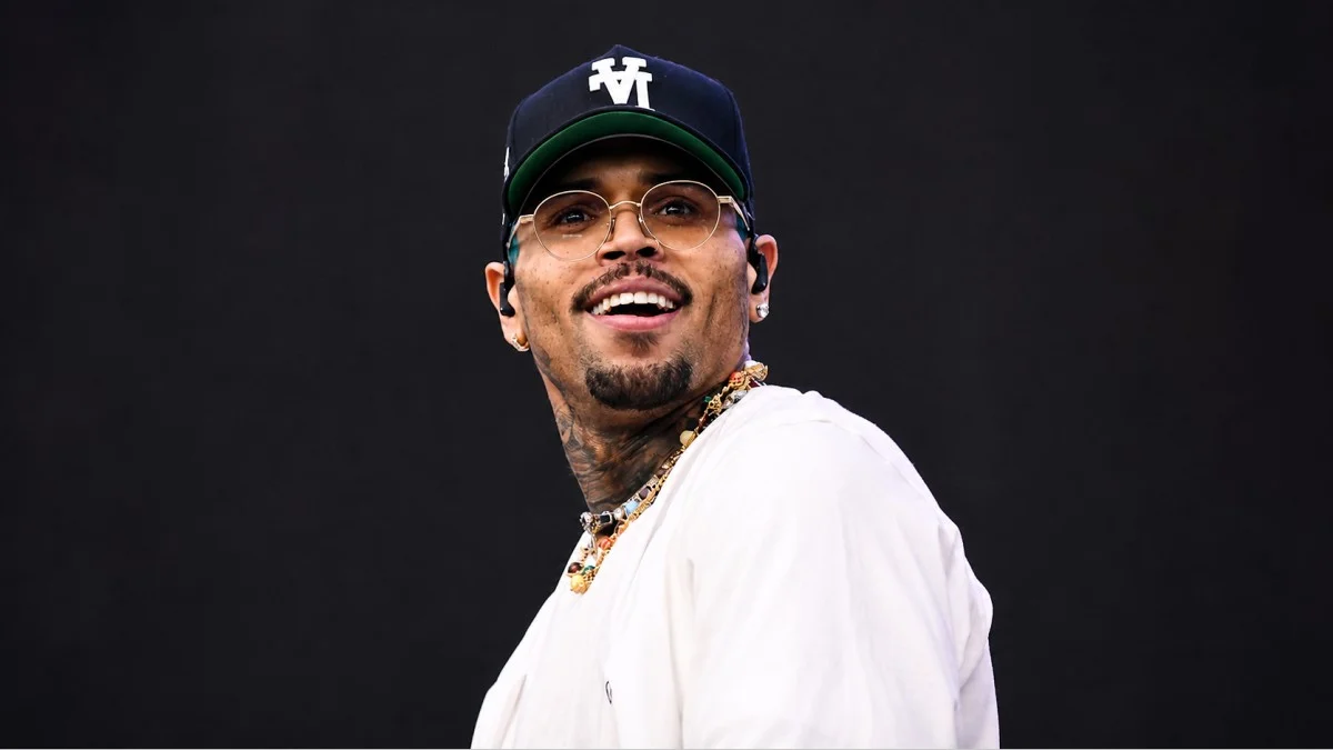 A close-up of Chris Brown smiling broadly on stage wearing a white shirt, black cap with green brim, round gold-framed glasses, layered gemstone necklaces, and neck tattoos visible — capturing the charismatic stage presence that has powered Chris Brown net worth to over $50 million throughout his career.