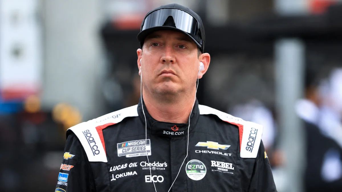 Kyle Busch wearing a professional racing suit with sponsor logos, standing in a motorsports setting with a focused expression.