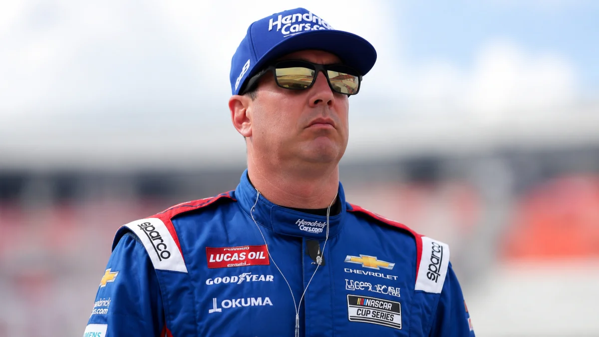 Kyle Busch wearing a blue racing cap and sunglasses while standing trackside in a professional racing suit with a focused expression.