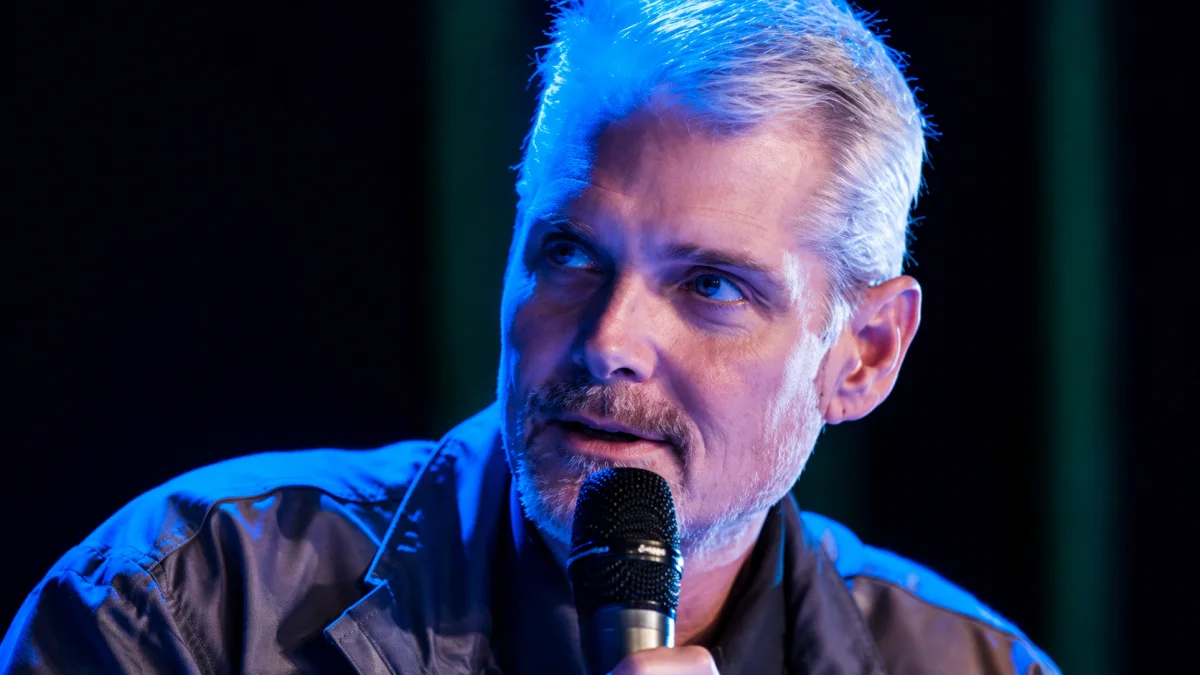 Tom Kane speaking into a microphone during a public event with blue stage lighting for Tom Kane biography and entertainment content.