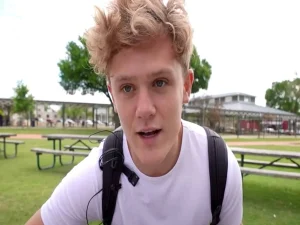 Ryan Trahan wearing a white t-shirt and black backpack speaking directly to camera outdoors in a park setting, in a style typical of his YouTube vlogs
