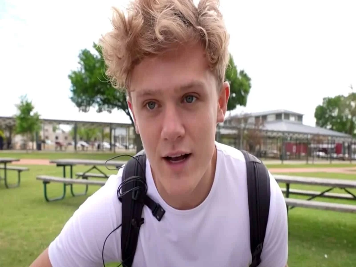 Ryan Trahan wearing a white t-shirt and black backpack speaking directly to camera outdoors in a park setting, in a style typical of his YouTube vlogs