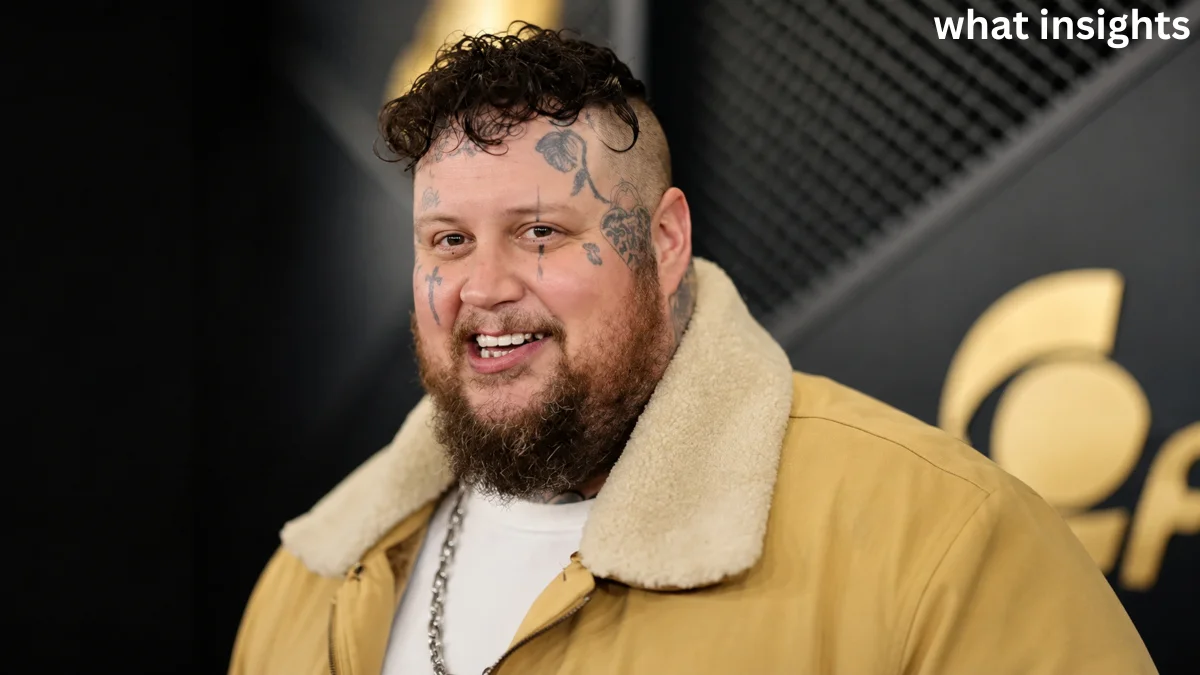 Jelly Roll smiling during a music industry event, wearing a tan jacket with visible facial tattoos and a beard.