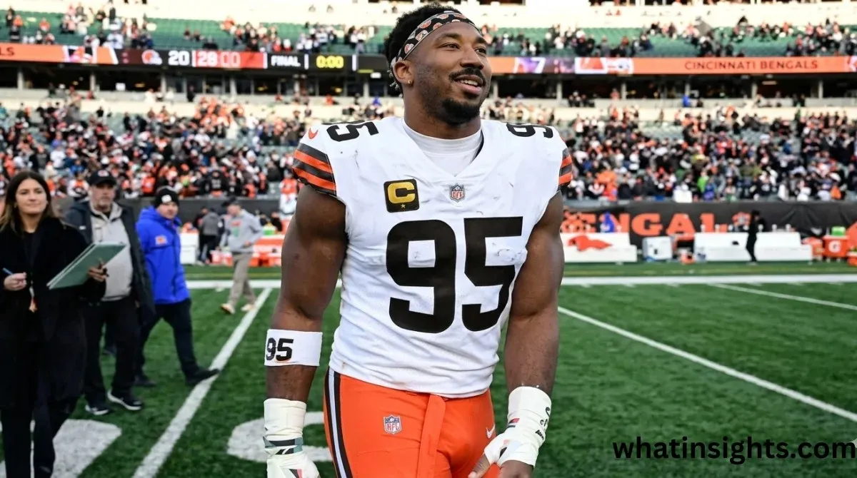 Cleveland Browns defensive end Myles Garrett walking on the football field in his white away jersey with number 95 and orange pants, smiling after a game with stadium fans in the background.